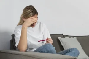 Mulher loira angustiada sentada no sofá, cobrindo o rosto com a mão e segurando um teste de gravidez, simbolizando a frustração e a luta para conceber, um desafio comum para quem enfrenta anovulação.