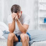 Homem sentado na cama com as mãos no rosto, preocupado com a relação entre torção testicular e fertilidade.