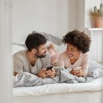 Um homem e uma mulher deitados na cama e pesquisando no celular se existem posições para engravidar.