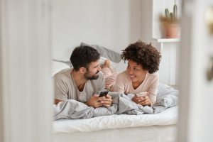 Um homem e uma mulher deitados na cama e pesquisando no celular se existem posições para engravidar.
