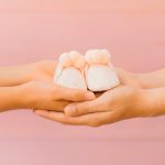 Mãos de uma mulher passando sapatinhos de bebê para outra, simbolizando a doação de embriões.