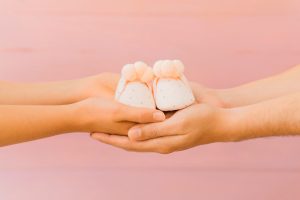 Mãos de uma mulher passando sapatinhos de bebê para outra, simbolizando a doação de embriões.
