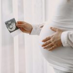 Mulher grávida com uma mão na barriga e a outra segurando a foto de um ultrassom para simbolização a implantação do embrião após a FIV.