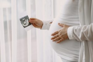 Mulher grávida com uma mão na barriga e a outra segurando a foto de um ultrassom para simbolização a implantação do embrião após a FIV.