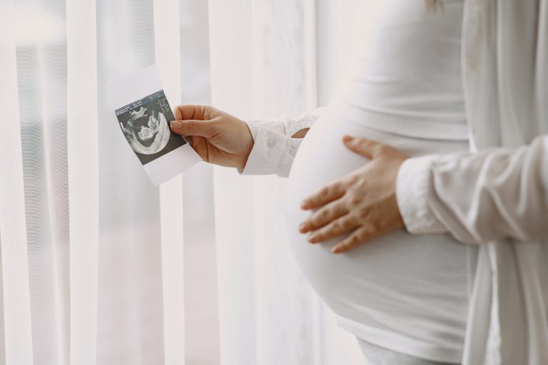 Mulher grávida com uma mão na barriga e a outra segurando a foto de um ultrassom para simbolização a implantação do embrião após a FIV.