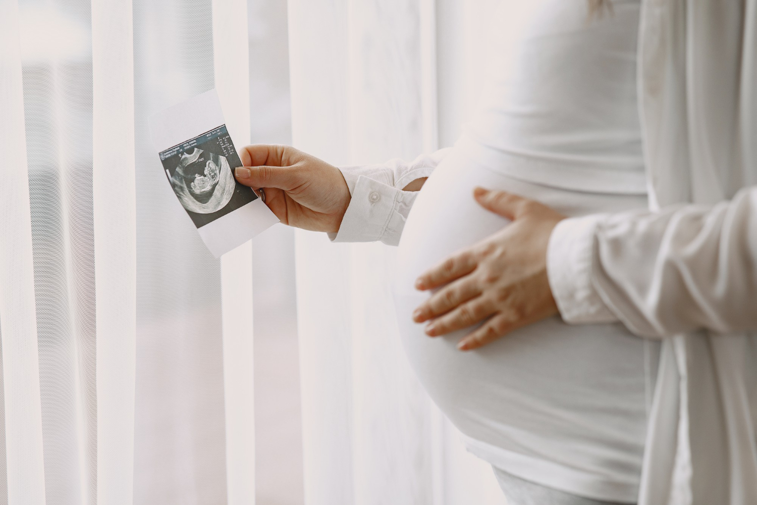 Mulher grávida com uma mão na barriga e a outra segurando a foto de um ultrassom para simbolização a implantação do embrião após a FIV.