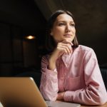 Mulher com a mão no queixo pensando "como saber se sou fértil?" em frente a um notebook para pesquisar sobre especialistas em fertilidade.