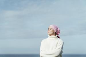 Portrait of young woman with cancer and crossed arms with closed eyes and sky on background